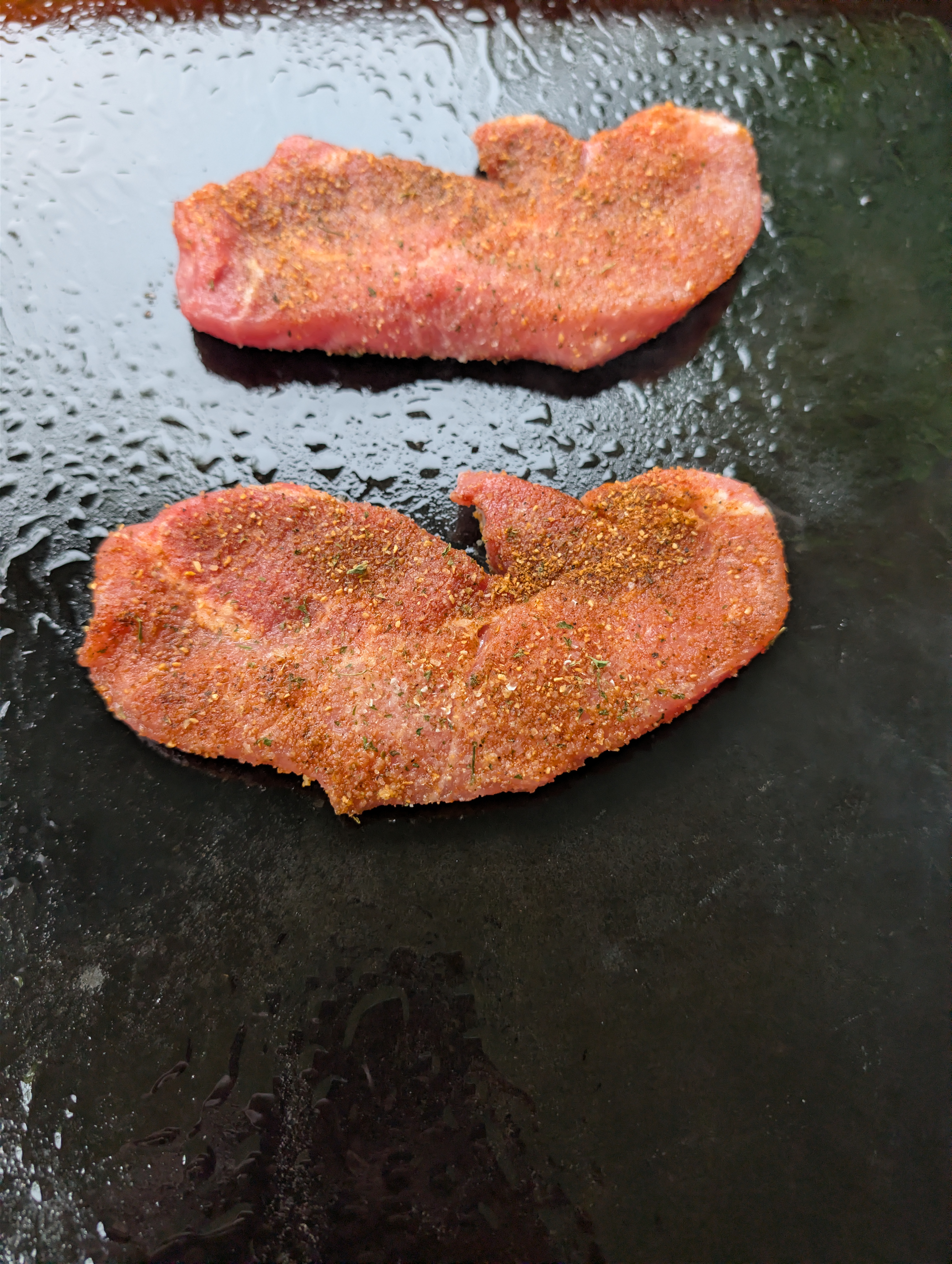 Seasoned pork steaks just placed on a hot Blackstone flat top griddle